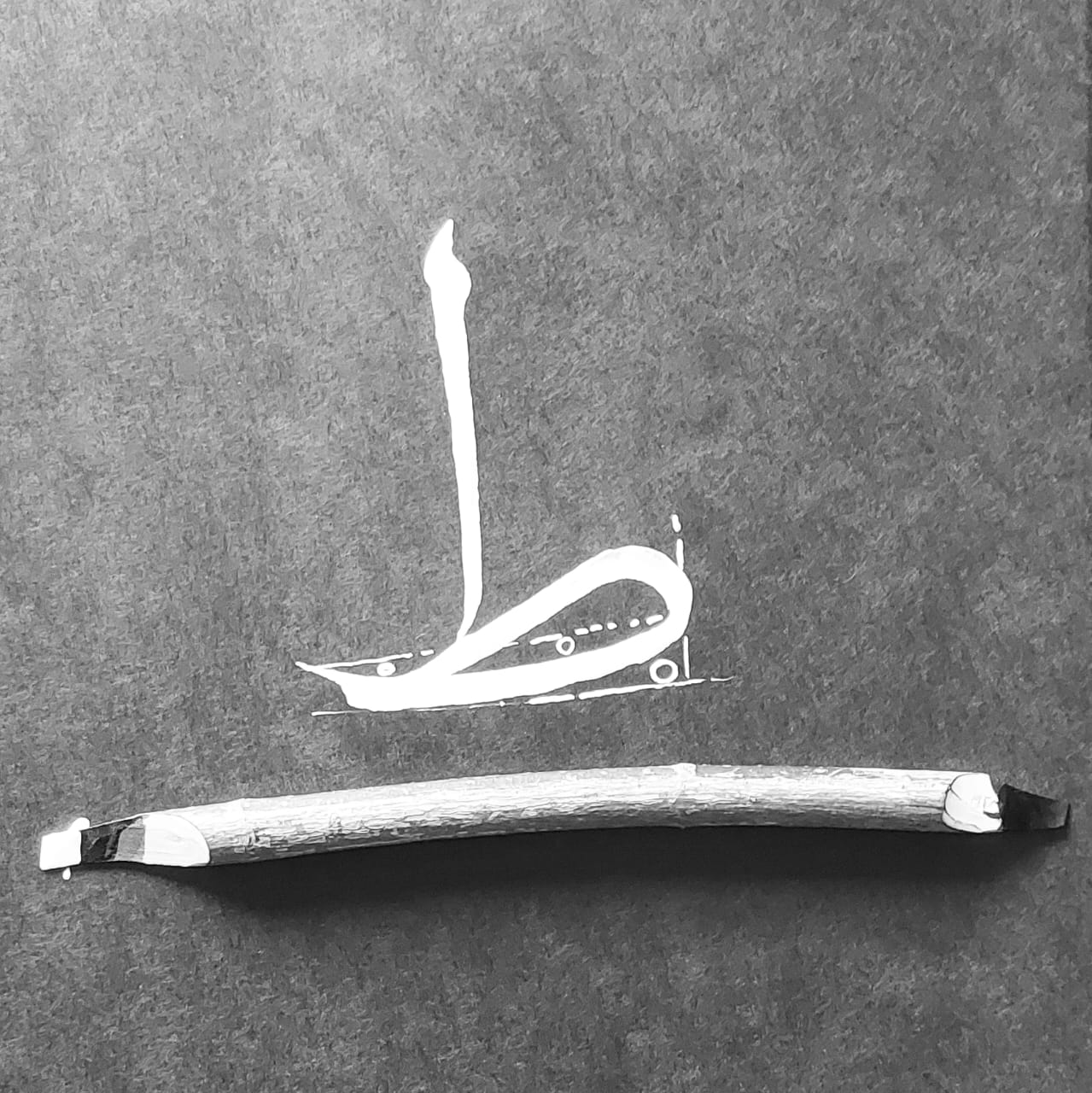 Image of the letter Taaw ط written in white ink on black paper with a Qalam below. 